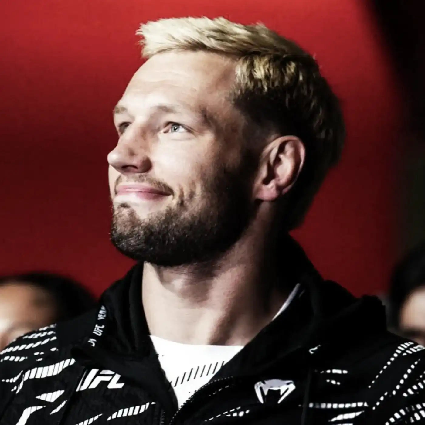 Blonde man met baard kijkt glimlachend opzij, gekleed in een sportjack met UFC-logo, tegen een rode achtergrond.