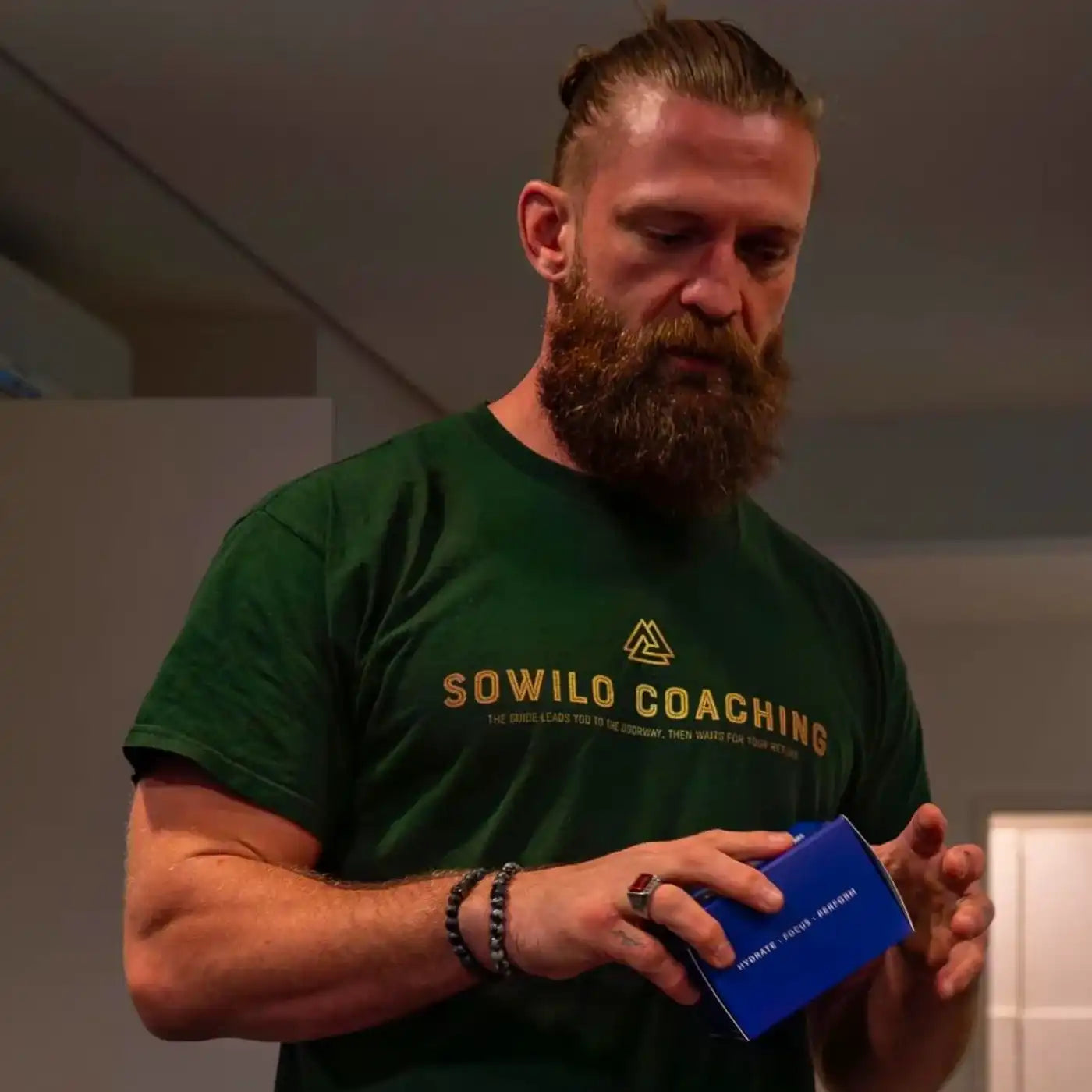 Bebaarde man in groen Sowilo Coaching-shirt houdt een blauw doosje vast en kijkt geconcentreerd naar beneden.