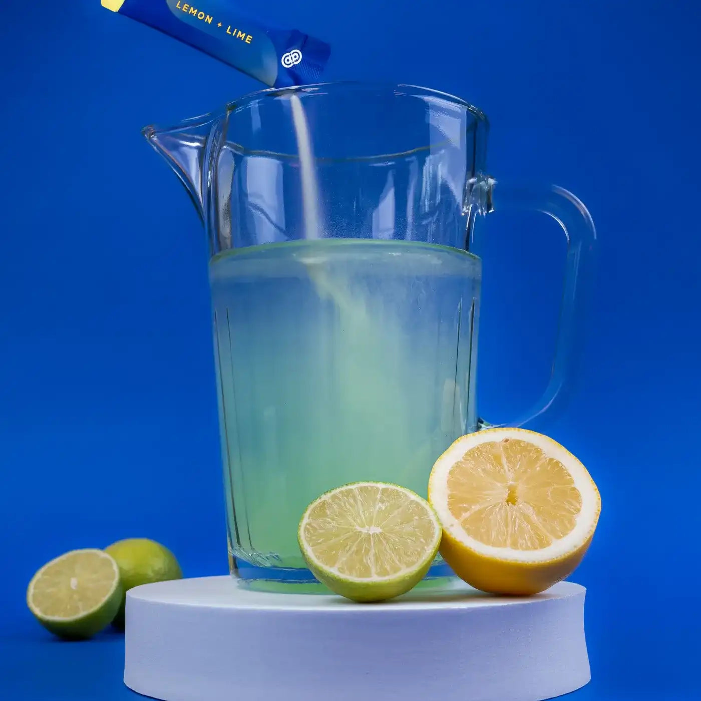 Glazen kan gevuld met lemon-lime drank, met een sachet dat wordt leeggegoten, en schijfjes citroen en limoen op een witte sokkel tegen een blauwe achtergrond.